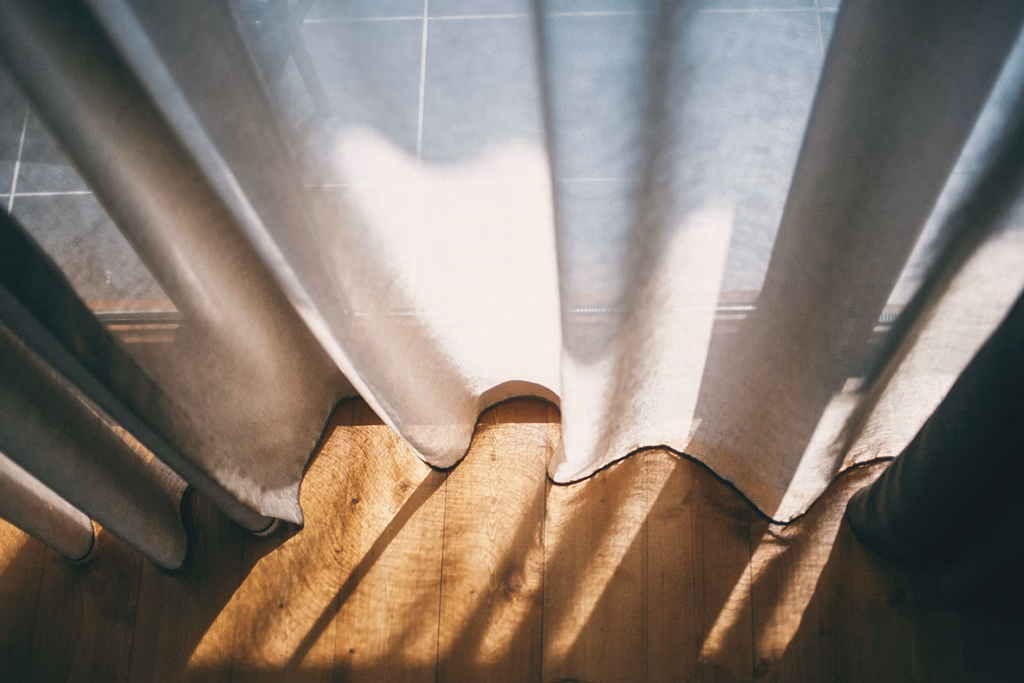 Un voilage de lin traversé par la lumière du matin, dont l'ombre se pose sur un parquet